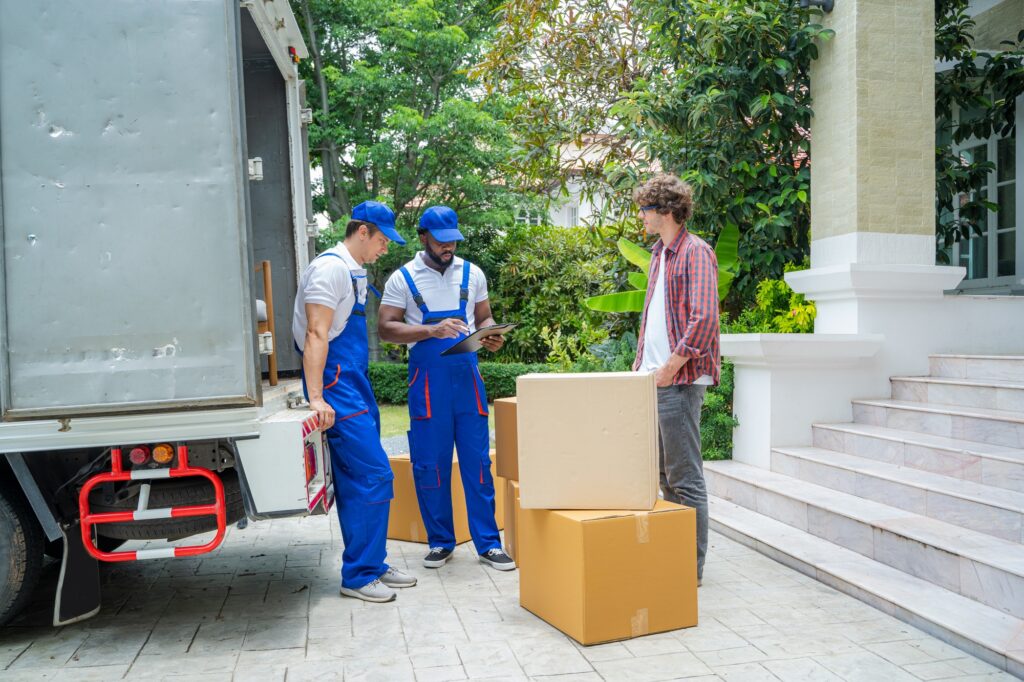 Deux déménageurs en vêtements bleus discutent avec un homme devant un camion, près de cartons empilés devant une maison.