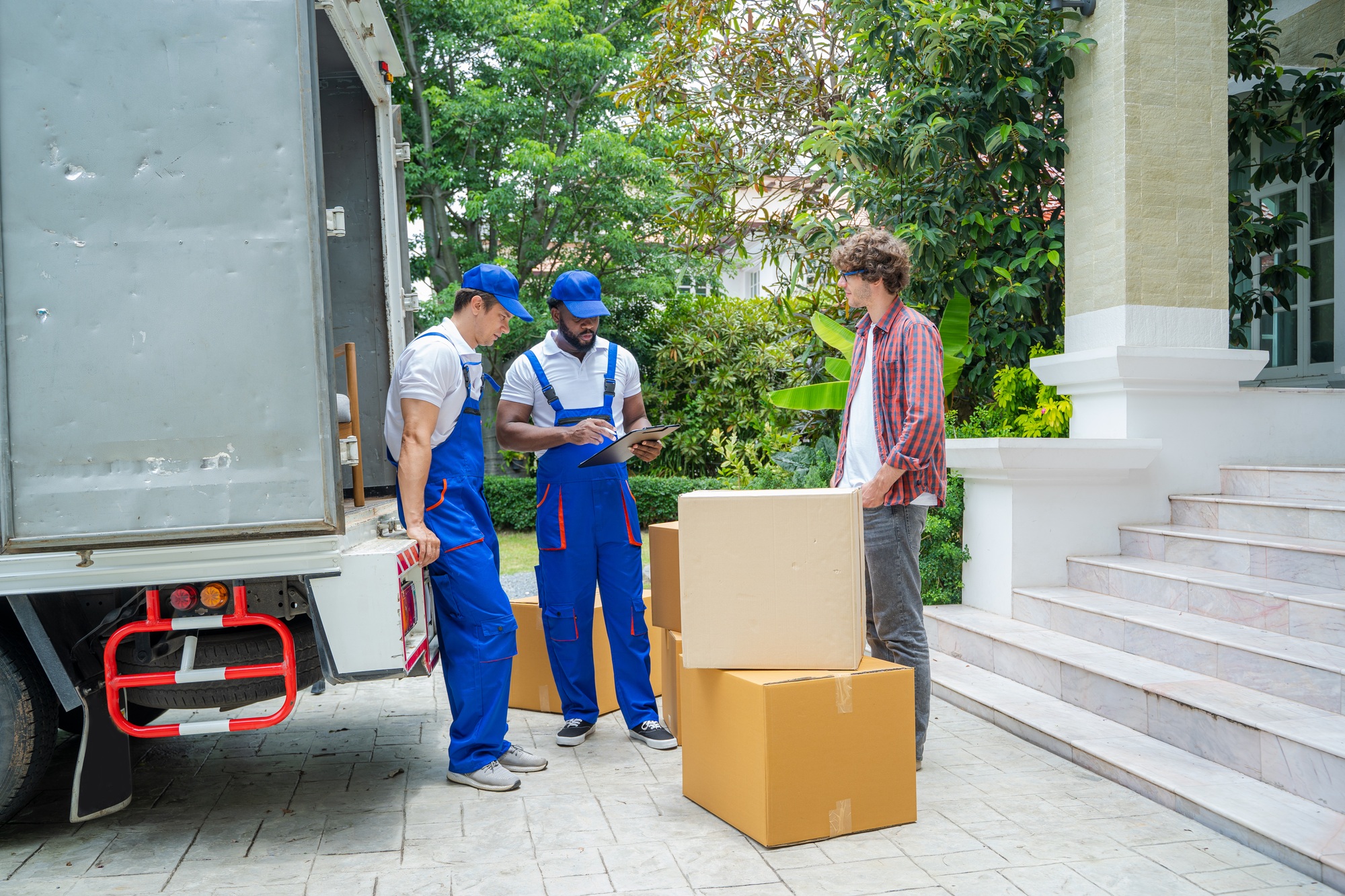 Deux déménageurs en vêtements bleus discutent avec un homme devant un camion, près de cartons empilés devant une maison.