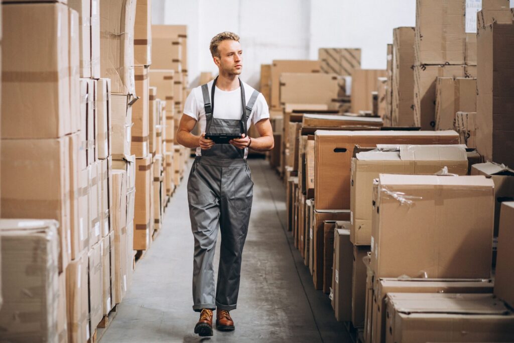 Livreur/ouvrier en entrepôt parmi des piles de cartons, tenant une tablette. intégrant le mot-clé checklist organiser son déménagement.